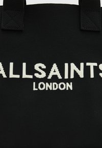 Fekete szövet bevásárlótáska, amelyen jól látható fehér "ALLSAINTS" és "LONDON" feliratok találhatók. A fogantyúk ugyanolyan anyagból készültek, minimalista dizájnnal.