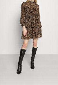 Robe imprimée animal marron avec de longues manches bouffantes, taille smockée, jupe fluide. Associée à de grandes bottes à talons brillantes noires.
