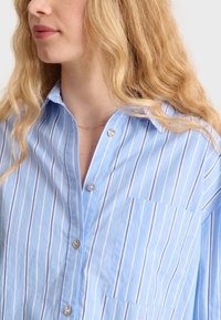 Camicia azzurra chiara con sottili righe verticali bianche, dotata di una sola tasca sul petto e bottoni simili a perle sulla patta.