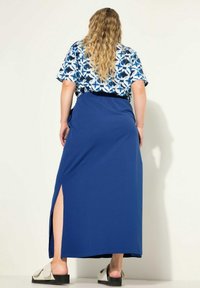 Maxi-jurk met een blauwe rok en een patroon in wit en blauw op de top. Heeft een zijsplit en een elastische tailleband. Gekozen in combinatie met platte sandalen.