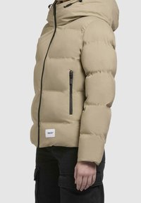 Beżowa pikowana kurtka z kapturem, z przodu zapinana na zamek błyskawiczny, z czarnymi akcentami i kieszenią boczną. Teksturowany materiał, zaprojektowany dla ciepła.