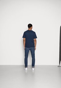 Mörkblå t-shirt i bomull med korta ärmar, i kombination med blå jeans och vita sneakers, sedd bakifrån mot en enfärgad bakgrund.