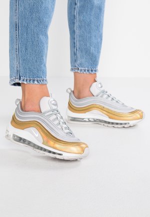 Person iført hvide og gyldne atletiske sneakers med synlige air-såler og lyseblå jeans mod en hvid baggrund.