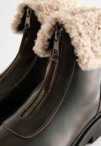 Brown leather boots with a fluffy beige shearling collar, featuring dual zippers on the sides and contrasting brown stitching details.
