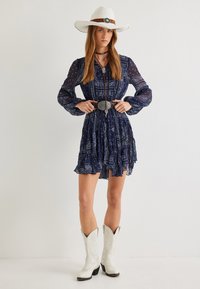 Laivansiniset mekko, jossa on pitkät hihat, röyhelöhelma ja paisley-kuvio, yhdistetty valkoisiin cowboy-kengiin ja valkoiseen hattuun, jossa on ruskea nauha.