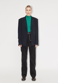 Blazer bleu marine surdimensionné sur un pull côtelé vert, associé à un pantalon noir texturé et des bottes foncées. Présente un détail en chaîne argentée sur la ceinture.
