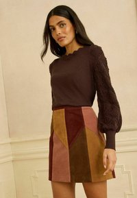 Pull en maille marron avec manches texturées, associé à une mini-jupe en daim multicolore présentant des motifs géométriques en rouille, moutarde et olive.
