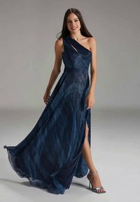 Mujer con un vestido camisón azul marino de un solo hombro y abertura, sonriendo y posando en sandalias de tacón plateadas contra un fondo plain.