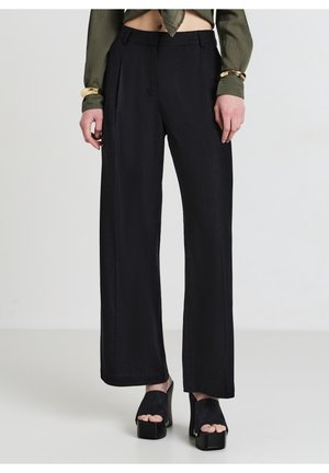 Personne portant un pantalon noir à jambes larges et des chaussures plateformes noires à bout ouvert, avec un haut vert court et des bracelets dorés aux poignets.