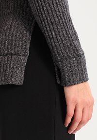 Šedý žebrovaný svetrový rukáv s texturovanou látkou, s bočním rozparkem a úzkým manžetou, v kombinaci s černým oděvem pod ním.