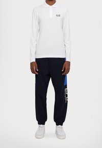 Vit långärmad polo med krage och EA7-logotyp, i kombination med marinblå träningbyxor med blå detaljer och logotryck; vita sneakers.