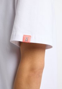 Witte katoenen t-shirt mouw met een roze label met een grafisch ontwerp. Gladde textuur en klassieke pasvorm. Labeldetail zichtbaar aan de rand van de mouw.