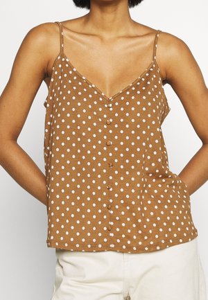 Bruine mouwloze top met witte stippen en met stof bedekte knopen, gedragen met lichte broek, handen achter de rug.