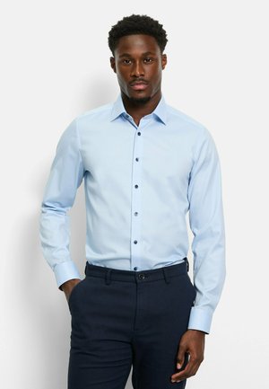 Hellblaues langärmliges Hemd mit Button-Down-Kragen, vertikaler Textur und dunklen Knöpfen. Kombiniert mit dunklen gemusterten Hosen.
