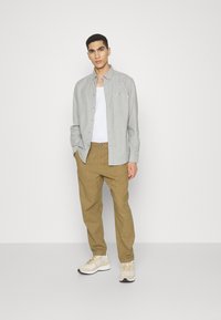 Camisa de botones gris claro sobre una camiseta blanca sin mangas, acompañada de pantalones beige y zapatillas color crema. Incluye un bolsillo y tiene un corte holgado.