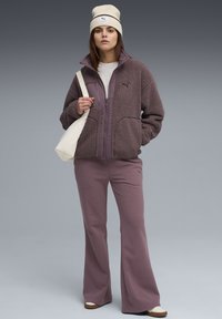 Giacca di pile viola con chiusura a zip e tasche frontali, abbinata a pantaloni viola coordinati e un berretto crema. Borsa a tracolla bianca.