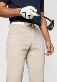 Μπεζ γκολφ παντελόνι με υφή, συνδυασμένο με μια λευκή ζώνη και μια ναυτική μπλούζα. Ένα λευκό γκολφ γάντι κρατά μια μαύρη γκολφ ράβδο.