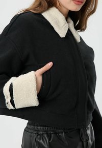 Zwarte cropped jas met een beige fleece kraag en manchetten. Voorzien van een zijzak en een verstelbare zoom met elastische details.