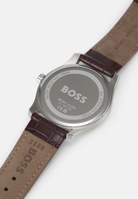 Reloj de acero inoxidable con esfera plateada, correa de cuero auténtico marrón y logo grabado. Presenta una caja resistente al agua.