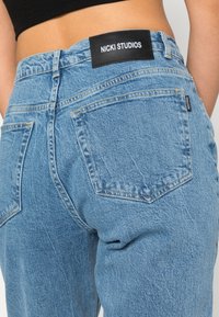 Jeans de denim azul claro com uma cintura alta, duas bolsas traseiras e uma etiqueta de couro preta com a marca "NICKI STUDIOS" na cintura.