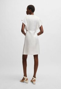 Biała sukienka z krótkimi falbaniastymi rękawami, strukturą materiału, dopasowaną talią i prostym krojem. Połączona z beżowymi sandałami na słupku.