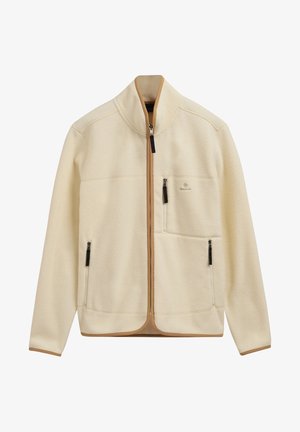 Veste polaire couleur crème avec fermeture éclair intégrale à l'avant, deux poches latérales, une poche poitrine, et bordure beige le long des bords.