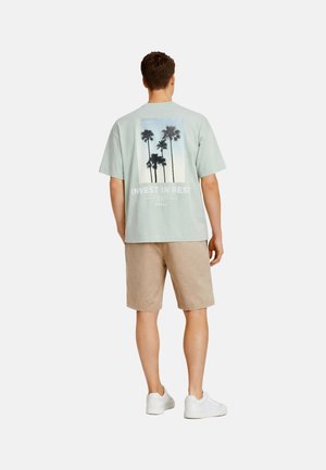 Man met een lichtgroen t-shirt met palmboomprint en de tekst "INVEST IN REST", beige korte broek en witte sneakers, staand met de rug naar de camera.