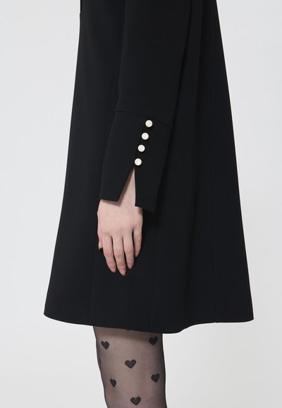 Schwarzes Kleid mit einem ausgestellten Design, das mit Knopfakzenten an den Ärmeln versehen ist. Kombiniert mit transparenten Strumpfhosen, die mit kleinen Herzen gemustert sind.
