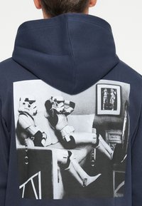Granatowy hoodie z dużą czarno-białą grafiką przedstawiającą dwóch szturmowców relaksujących się na kanapie, otoczonych czarną ramką na ścianie.