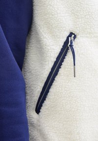 Fleece striukė su balta, tekstūruota kūno dalimi ir tamsiai mėlynomis rankovėmis, su užtrauktuko kišene ir sidabriniu trauktuku.