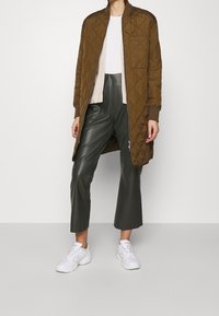 Brauner, gesteppten langer Mantel mit gerippten Bündchen, kombiniert mit einem cremefarbenen Pullover, dazu dunkelgrüne, kurz geschnittene Hose aus Kunstleder und weiße Sneaker.