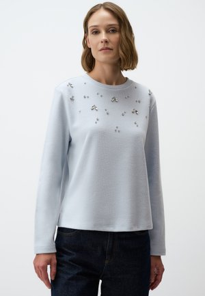 Maglione azzurro chiaro a maniche lunghe con finitura testurizzata e decorazioni argento sulla parte frontale, abbinato a jeans in denim scuro.
