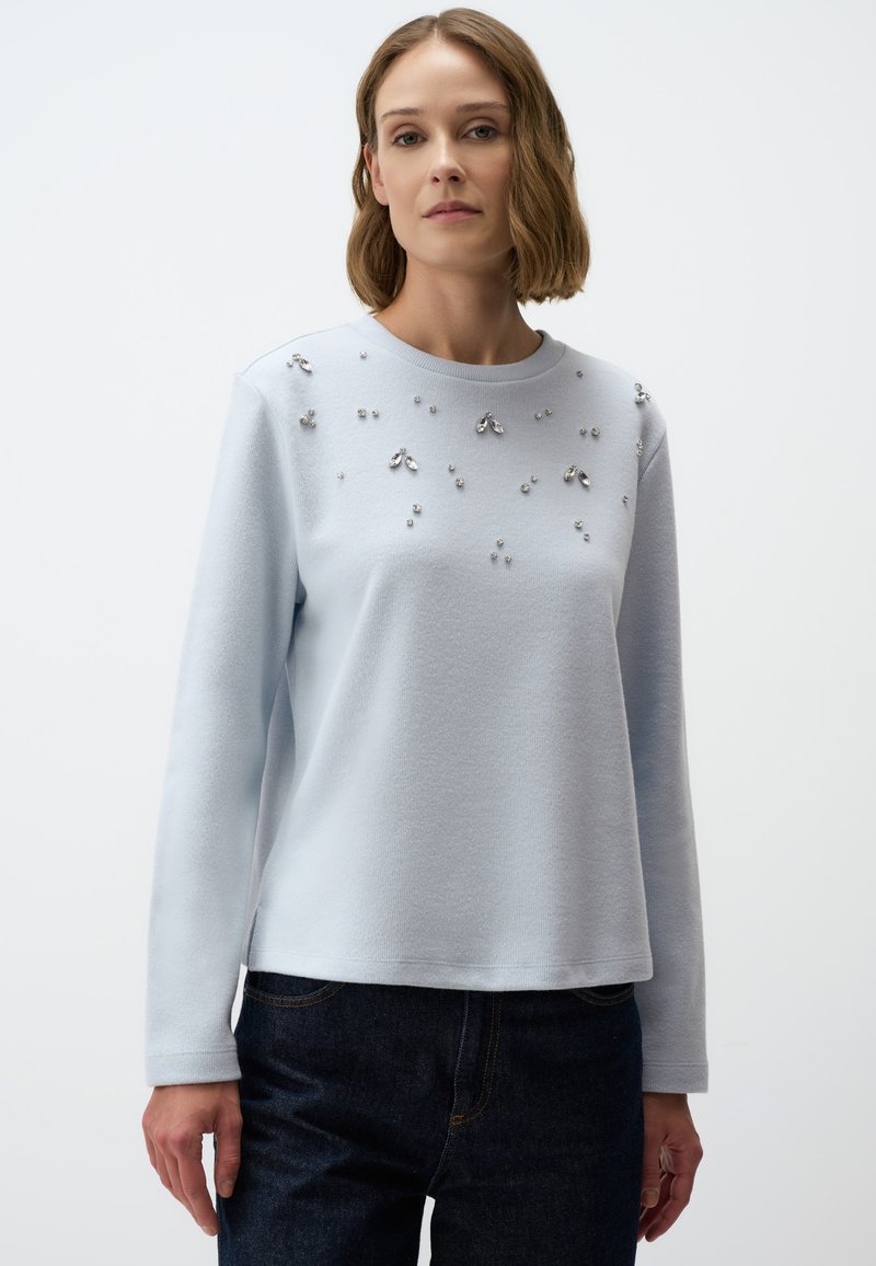 Hellblauem Pullover mit langen Ärmeln, strukturiertem Finish und dekorativen silbernen Verzierungen auf der Vorderseite, kombiniert mit dunkelblauen Denim-Jeans.
