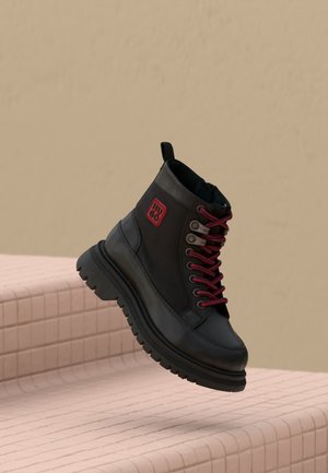 Stivale in pelle nera con lacci rossi e toppa del logo rossa che fluttua sopra gradini piastrellati rosa su uno sfondo beige.