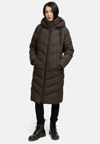 Cappotto imbottito marrone con cappuccio, dotato di chiusura con zip, trapuntatura a zigzag e tasche laterali, abbinato a pantaloni neri e stivali con lacci.