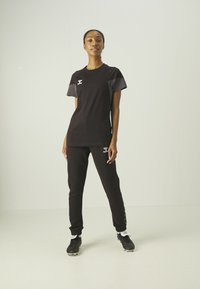 Maglietta sportiva nera con dettagli grigi e pantaloni neri coordinati con un logo e modelli testurizzati sui lati, abbinati a sneakers nere.