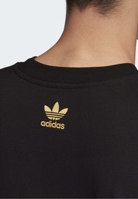 Svart sweatshirt med rundad halsringning, som har en gul Adidas-logotyp och bladdesign broderat på baksidan nära halsringningen. Mjuk textur.