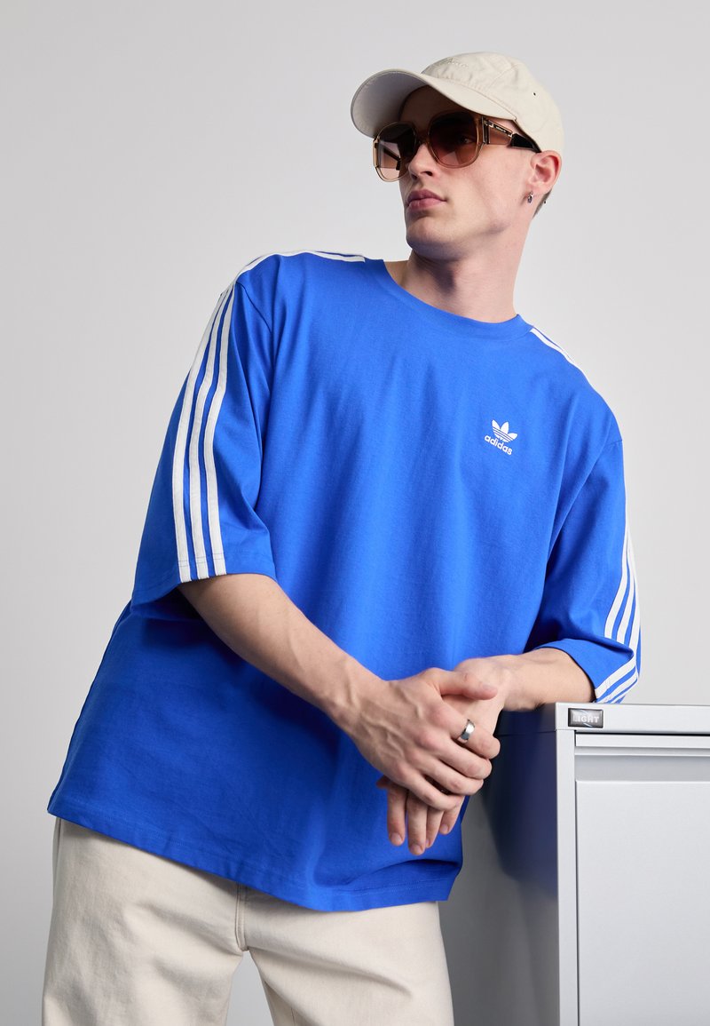 Kék, oversized pamut póló fehér Adidas logóval és három csíkkal az ujjakon. Férfi bézs sapkát visel és nagy napszemüveget, egy szürke szekrényre támaszkodik.