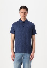 Polo azul-marinho com um padrão texturizado, placket de três botões e logotipo bordado no peito, combinado com jeans azul-claro.