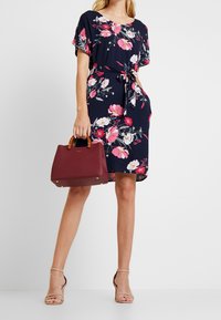 Marineblaues, blumiges Kleid mit kurzen Ärmeln und Taillengürtel. Das Modell hält eine bordeauxfarbene Handtasche mit braunen Griffen, kombiniert mit beigen Sandalen mit Riemchen.