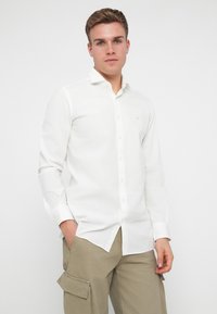 Hvit skjorte med krage, som har knappestolpe foran, langermet og en liten lyseblå logo på brystet, laget av glatt stoff.