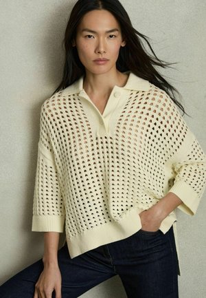 Maglione lavorato a maglia color crema con motivo a traforo, maniche corte e colletto a risvolto. Abbinato a jeans scuri. Tessuto leggero e strutturato.