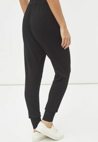 Pantalones de chándal negros con cintura elástica, puños ajustados y detalle de costura lateral. Combinados con zapatillas blancas sobre un fondo liso.