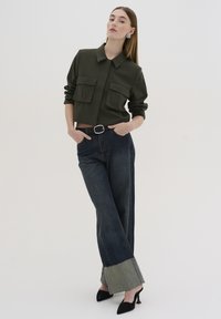 Chemise en olive courte avec deux poches avant, associée à un pantalon large bleu foncé avec un bas contrastant et des talons noirs.