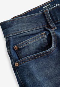 Jeans de mezclilla azul oscuro con un bolsillo estándar, herrajes de bronce y costuras en naranja contrastantes. Textura suave con un ligero desgaste.