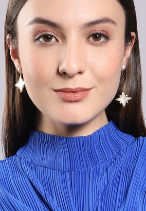 Goldfarbene Sternohrringe mit weißen Perlenakzenten, hängend unter einem unregelmäßigen Fransenbesatz, ergänzt durch ein blaues, plissiertes Oberteil.