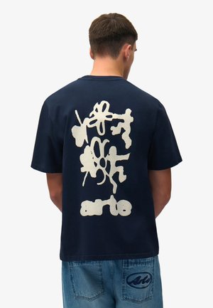 Uomo di spalle che indossa una maglietta blu navy con grafica astratta bianca e testo "arte", abbinata a pantaloncini di jeans blu con toppa sulla tasca.