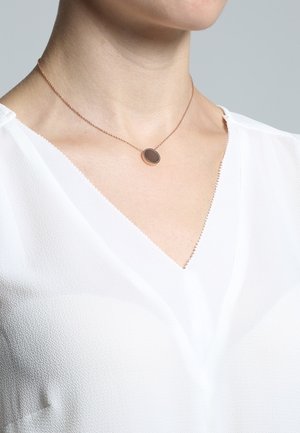 Femme portant un délicat collier chaîne en or rose avec un pendentif circulaire foncé, vêtue d'un chemisier blanc texturé à col en V.