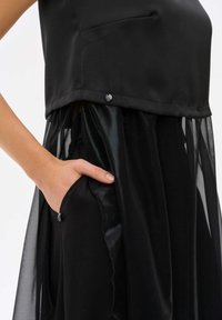 Zwarte gelaagde jurk met een satijnen bovenstuk met een zak en een sheer, vloeiende rok. Aangekleed met een decoratieve knoopdetail.