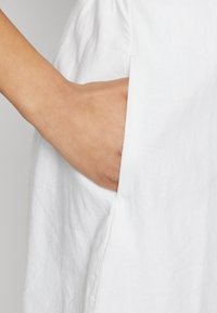 Λευκό λινό ένδυμα με πλευρική τσέπη. Η υφή φαίνεται ελαφρώς τσαλακωμένη, αναδεικνύοντας τις φυσικές ίνες του υφάσματος.